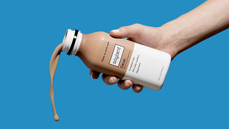 A bottle of Soylent Drink Original next to a backpack.