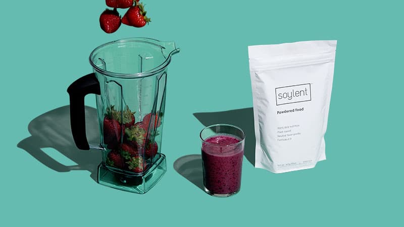 Blender with strawberries next to a Soylent Powder Pouch and a glass of smoothie.